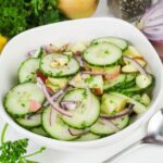 A white bowl filled with a salad of sliced cucumbers, red onions, apple pieces, and chopped herbs on a white surface.