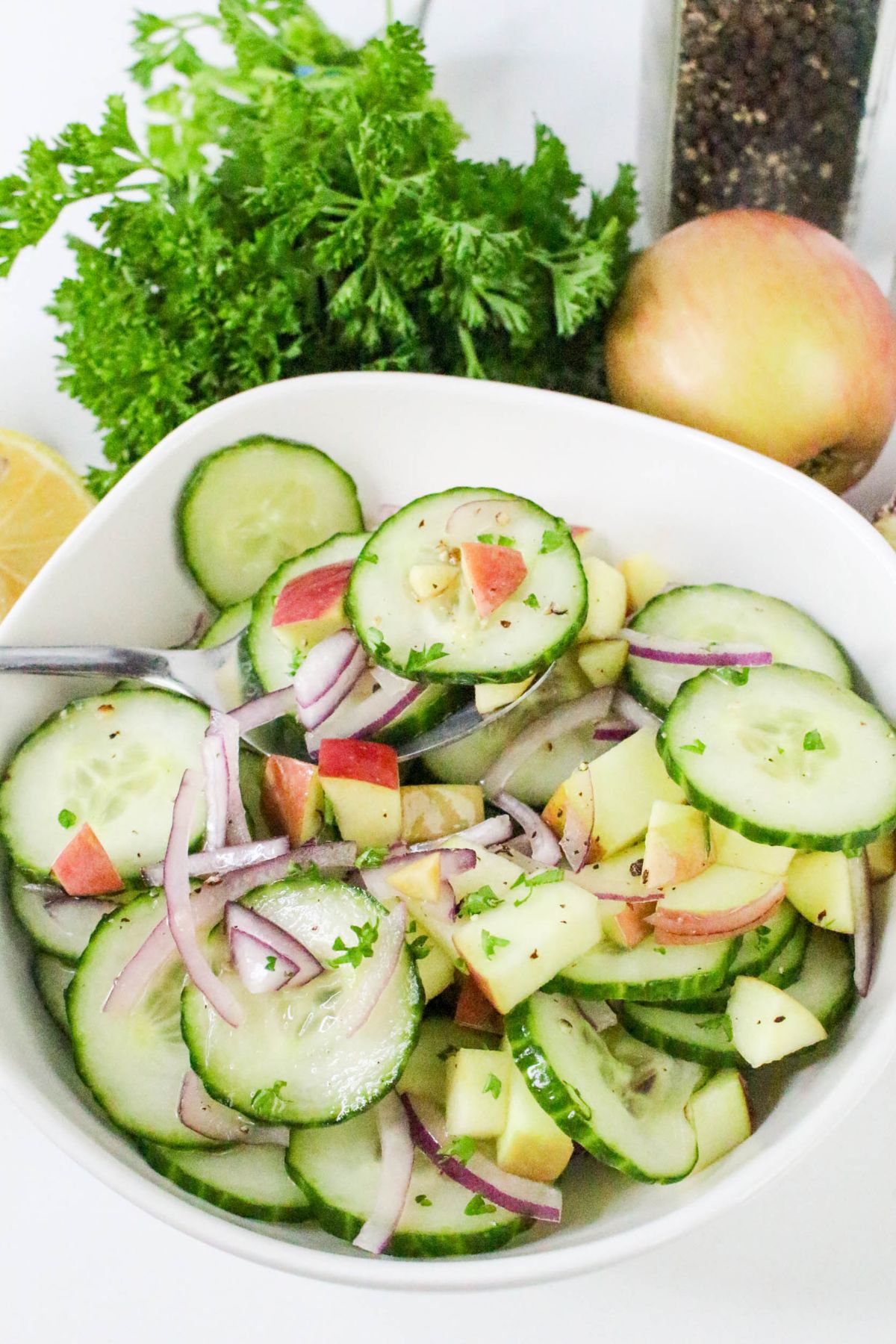 A bowl of cucumber salad with sliced cucumbers, red onion, apple pieces, and herbs, with a spoon, apple, lemon, parsley, and pepper grinder in the background.