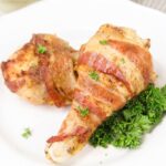 Two bacon-wrapped chicken drumsticks on a white plate, garnished with fresh parsley.