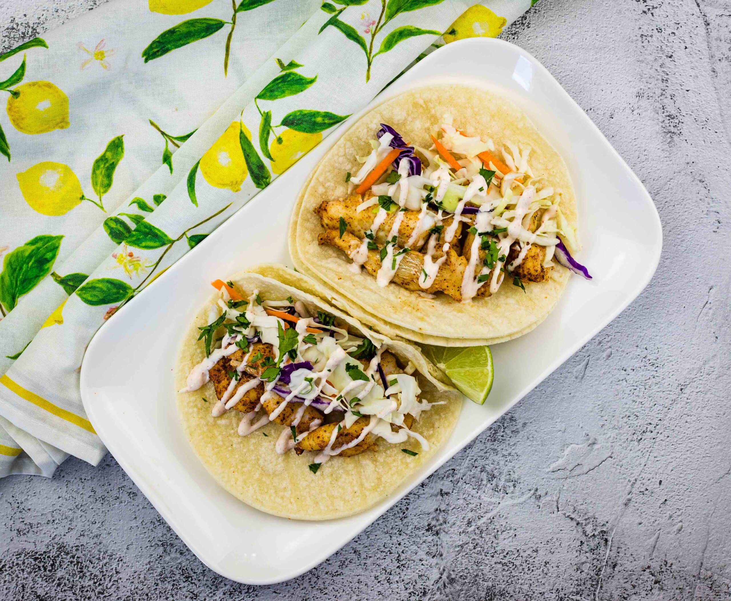 Two fish tacos with shredded cabbage and drizzled sauce on corn tortillas, served on a white plate with a lime wedge, next to a lemon-patterned napkin.