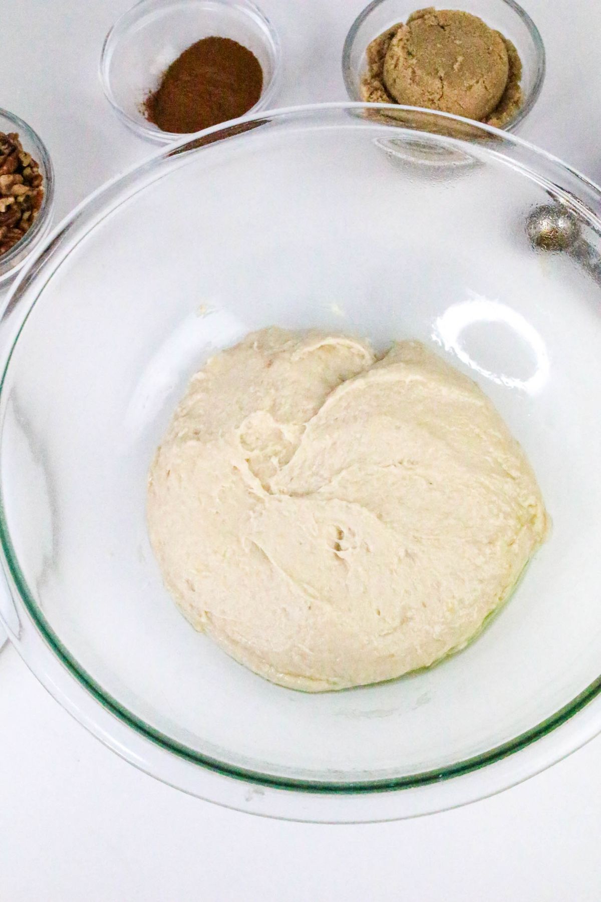 A glass bowl with dough in the center, surrounded by small bowls containing brown sugar, cinnamon, and chopped nuts on a white surface.