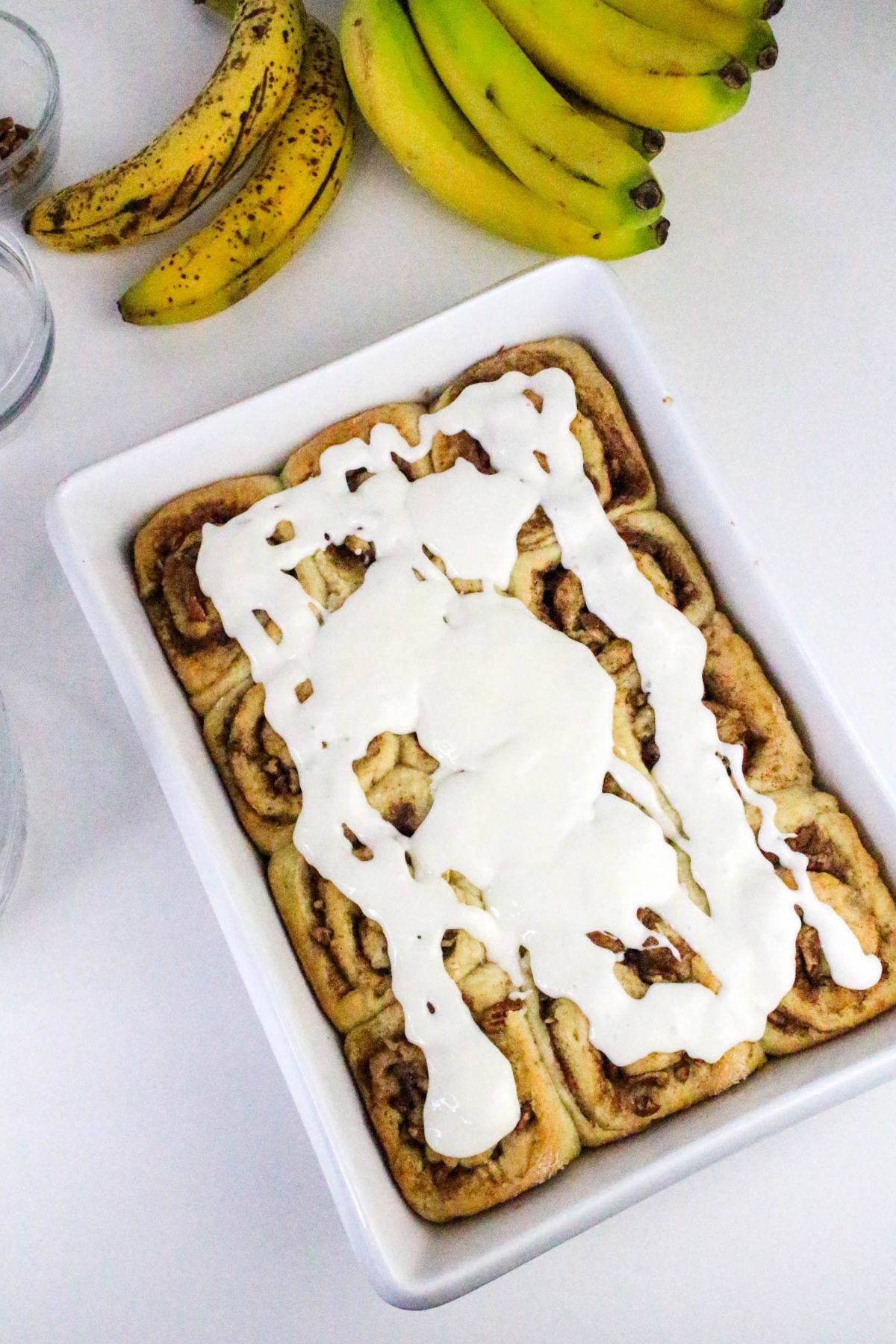 A white baking dish filled with cinnamon rolls topped with white icing, next to ripe and unripe bananas on a white surface.