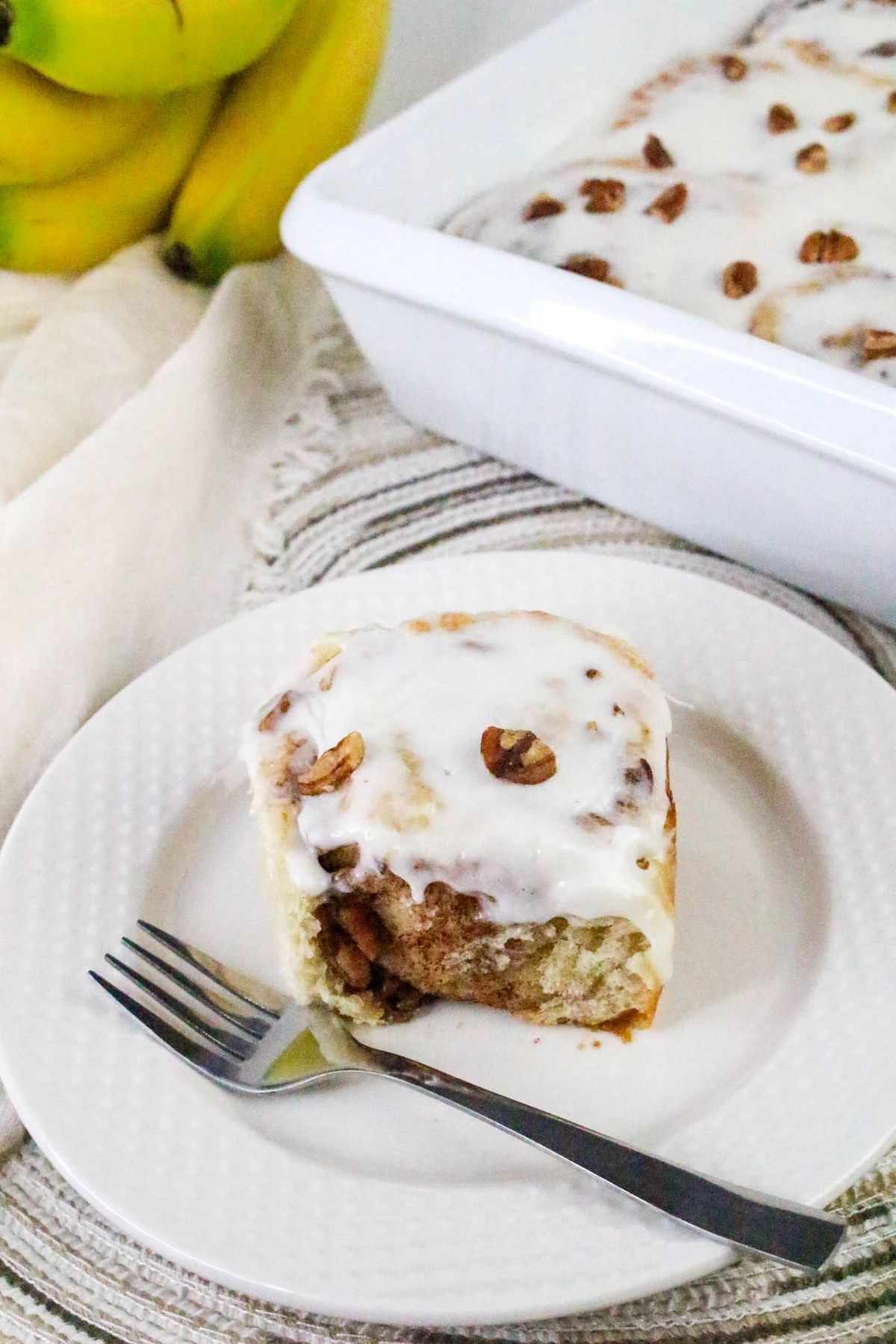 A frosted cinnamon roll topped with pecans on a white plate with a fork, next to a baking dish of more rolls and a bunch of bananas in the background.