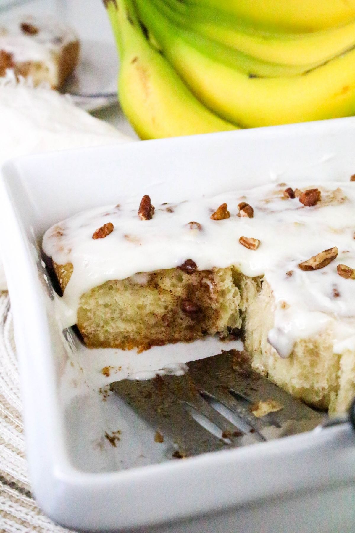 A frosted cinnamon roll with chopped pecans in a white baking dish, partially served, with bananas in the background.