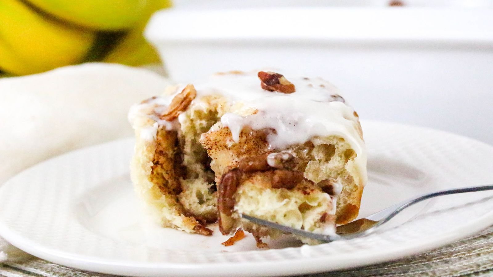 A cinnamon roll with icing and pecans on a white plate, partially eaten with a fork, with bananas and a cloth visible in the background.