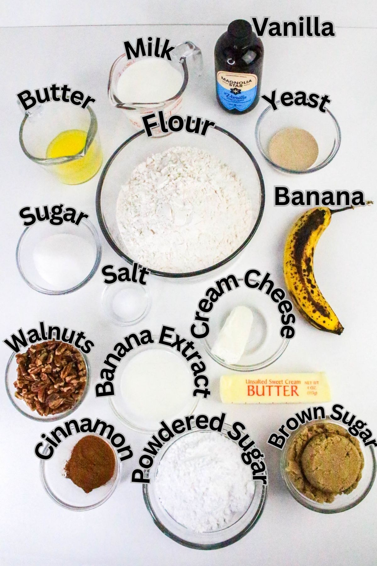 Ingredients for banana bread are arranged on a white surface, each labeled: flour, milk, vanilla, yeast, banana, cream cheese, butter, sugars, walnuts, salt, cinnamon, and banana extract.