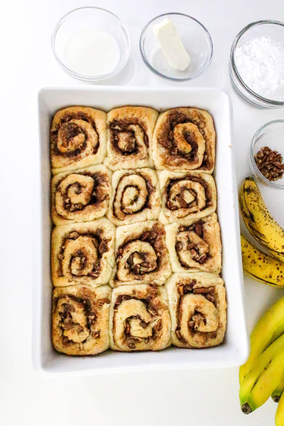 A white baking dish filled with twelve cinnamon rolls sits on a white surface, surrounded by bananas, pecans, powdered sugar, butter, and a small bowl of milk.