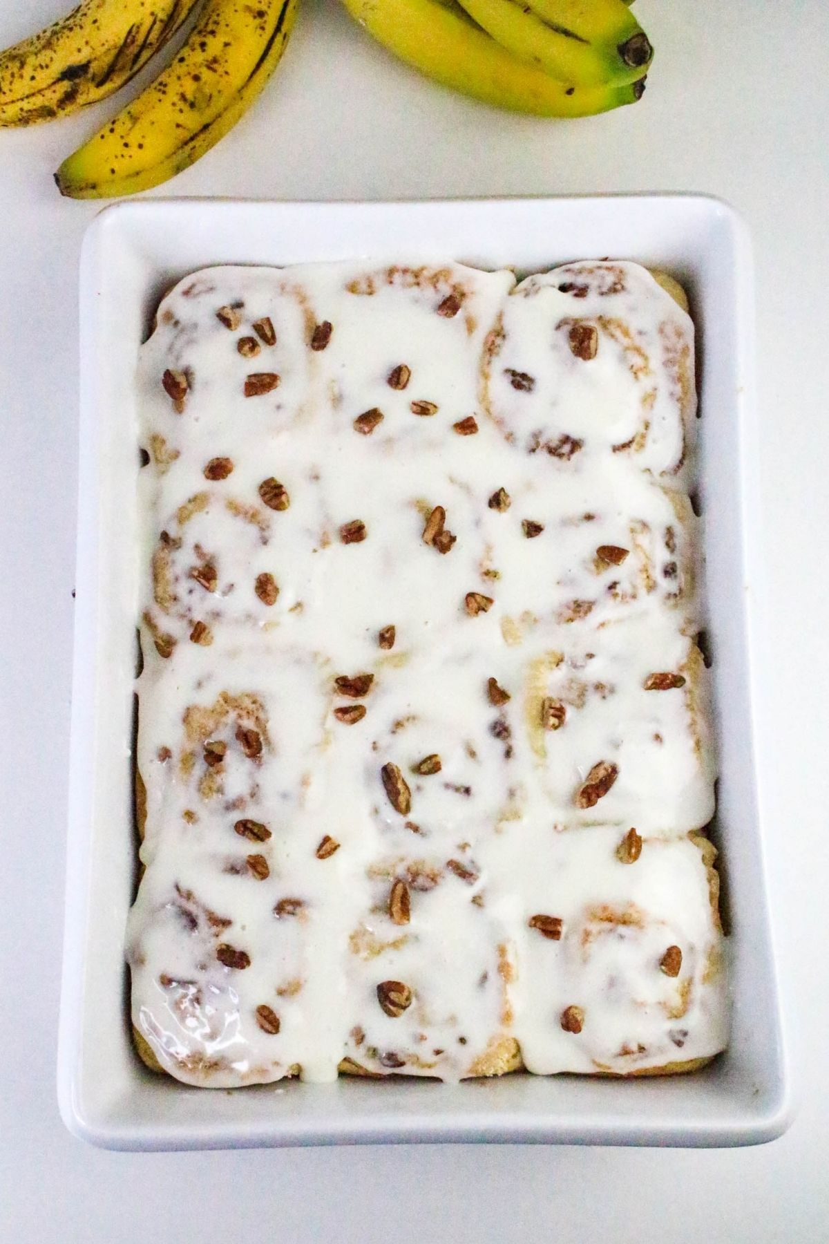 A rectangular dish filled with cinnamon rolls topped with white icing and scattered pecan pieces, with bananas in the background.
