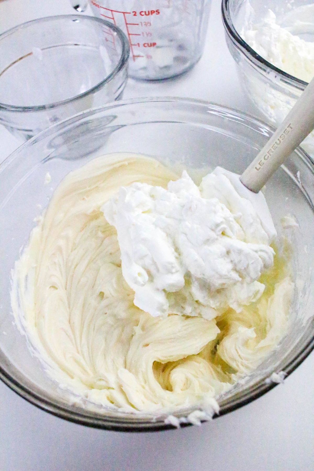 A clear mixing bowl with whipped cream being folded into a creamy mixture using a white spatula; other empty bowls are in the background.