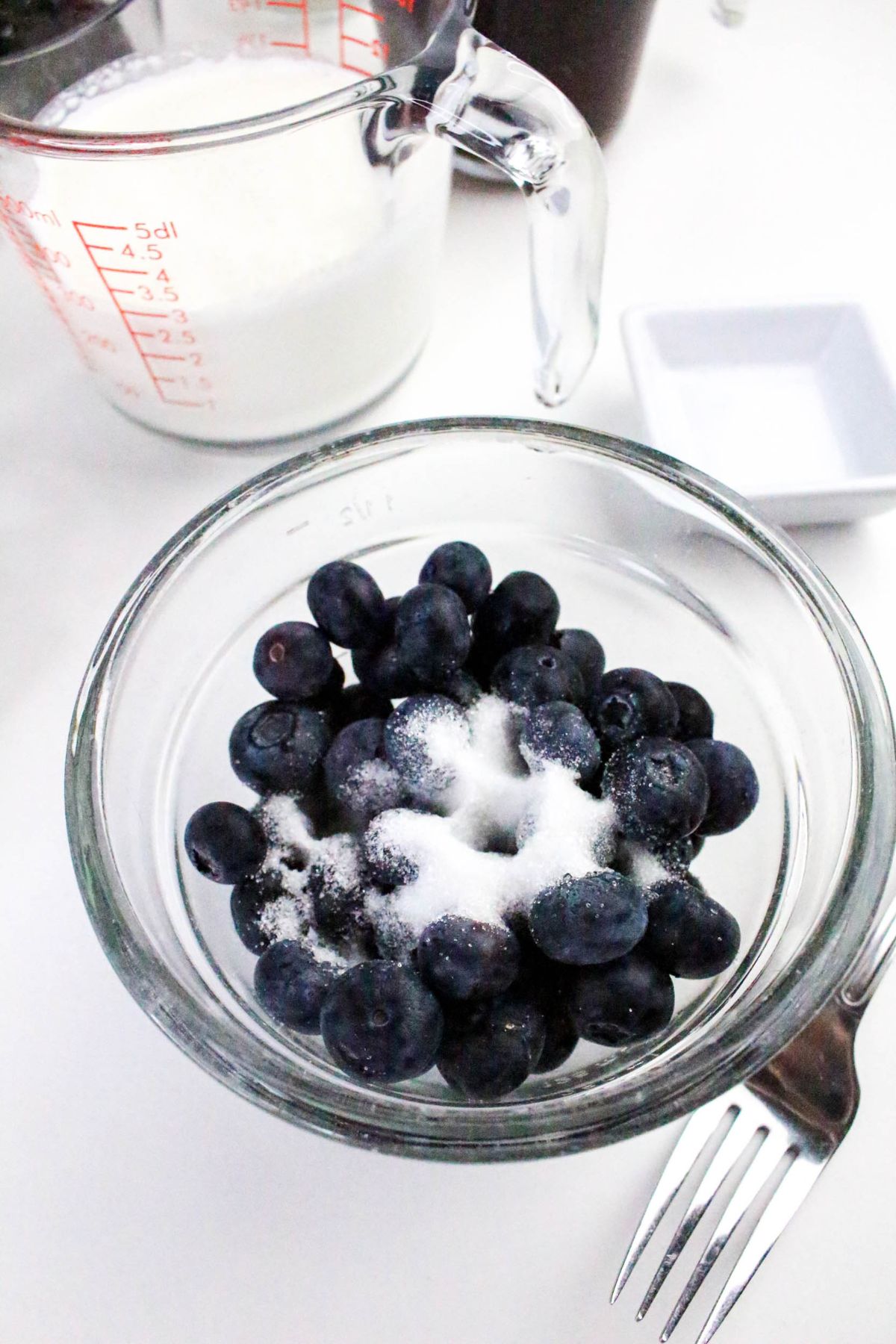 A glass bowl of blueberries sprinkled with sugar sits next to a fork, a measuring cup of milk, and a small white dish on a white surface.