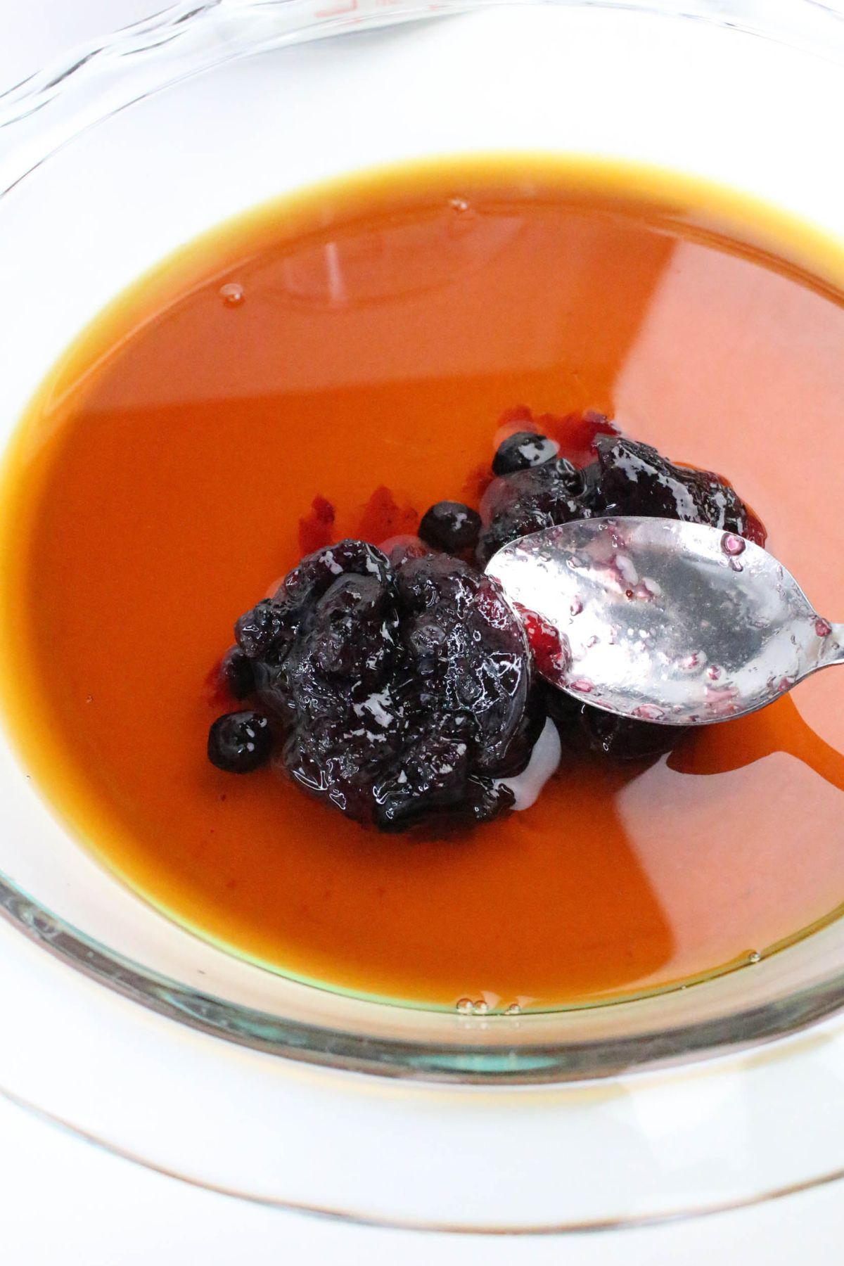 A silver spoon holds a dollop of dark berry preserves above a bowl of amber-colored liquid.