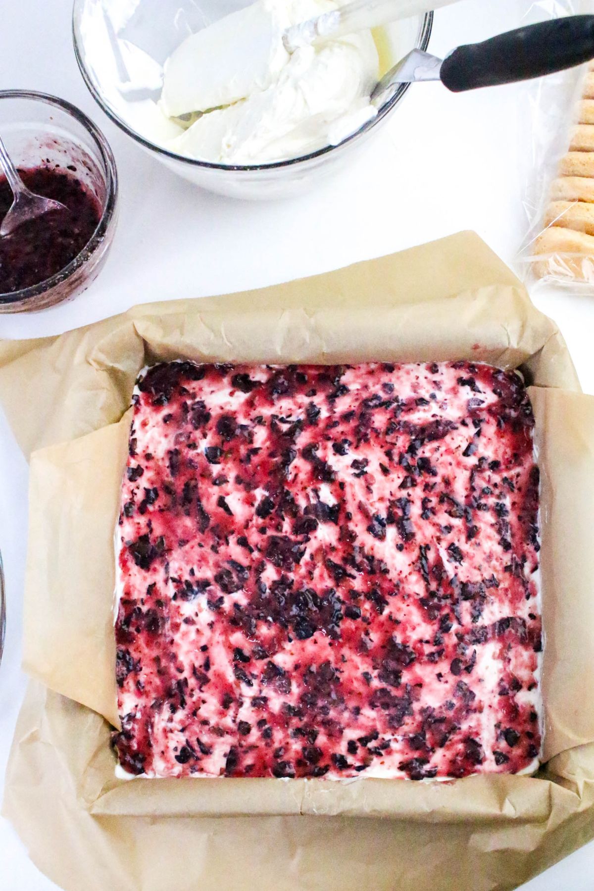 A square baking pan lined with parchment paper holds a marbled cheesecake mixture. Nearby are a bowl of cream, a jar of berry sauce, and a package of ladyfinger cookies.