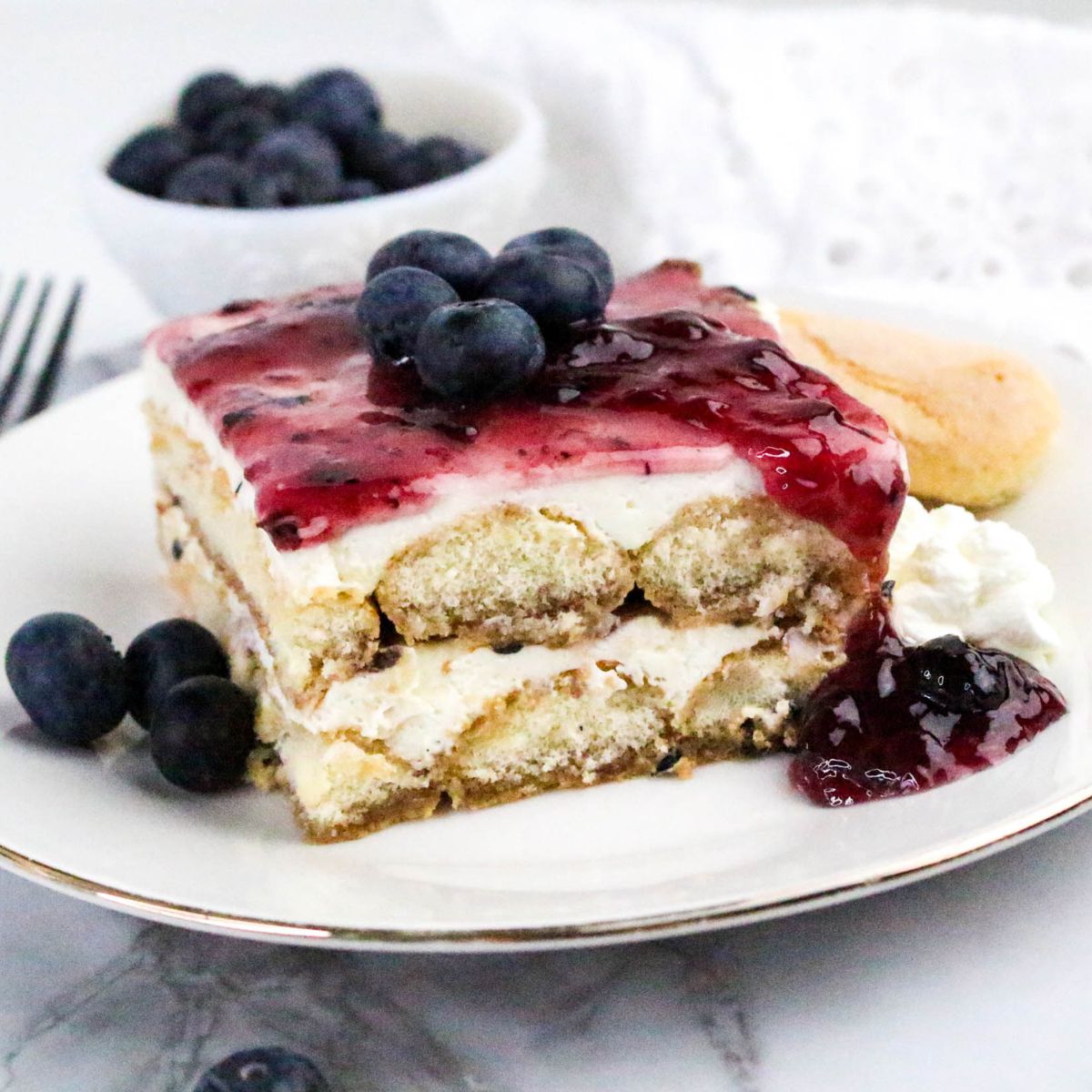 A slice of layered dessert topped with blueberry sauce and fresh blueberries, served on a white plate with whipped cream and extra blueberries on the side.