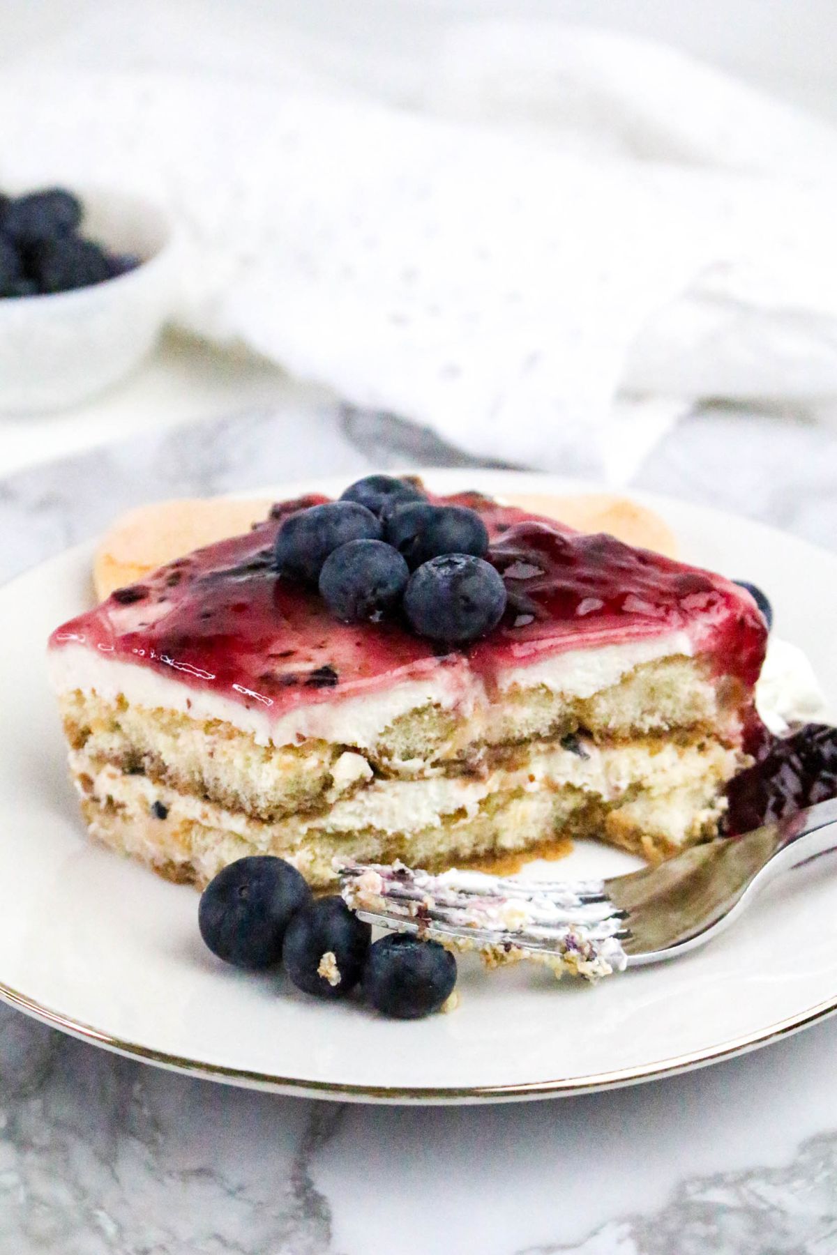 A slice of layered dessert topped with blueberry sauce and fresh blueberries is served on a white plate with a fork, alongside a few loose blueberries.