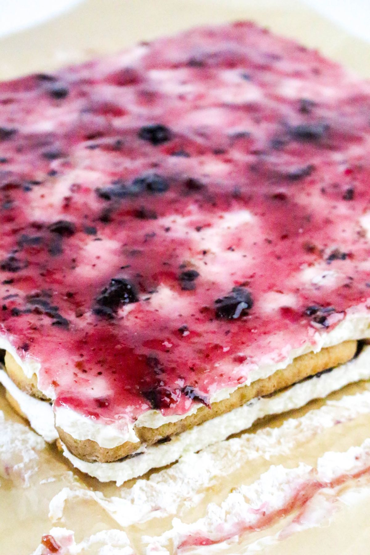 A rectangular dessert with a layer of cream filling and a glossy berry spread on top, partially covered with parchment paper.