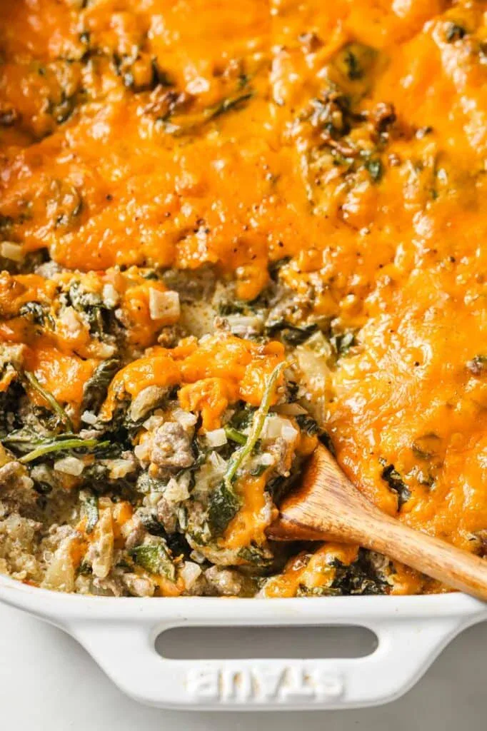 A close-up of a baked casserole with melted cheddar cheese, visible greens, and a wooden spoon scooping a portion from a white baking dish.
