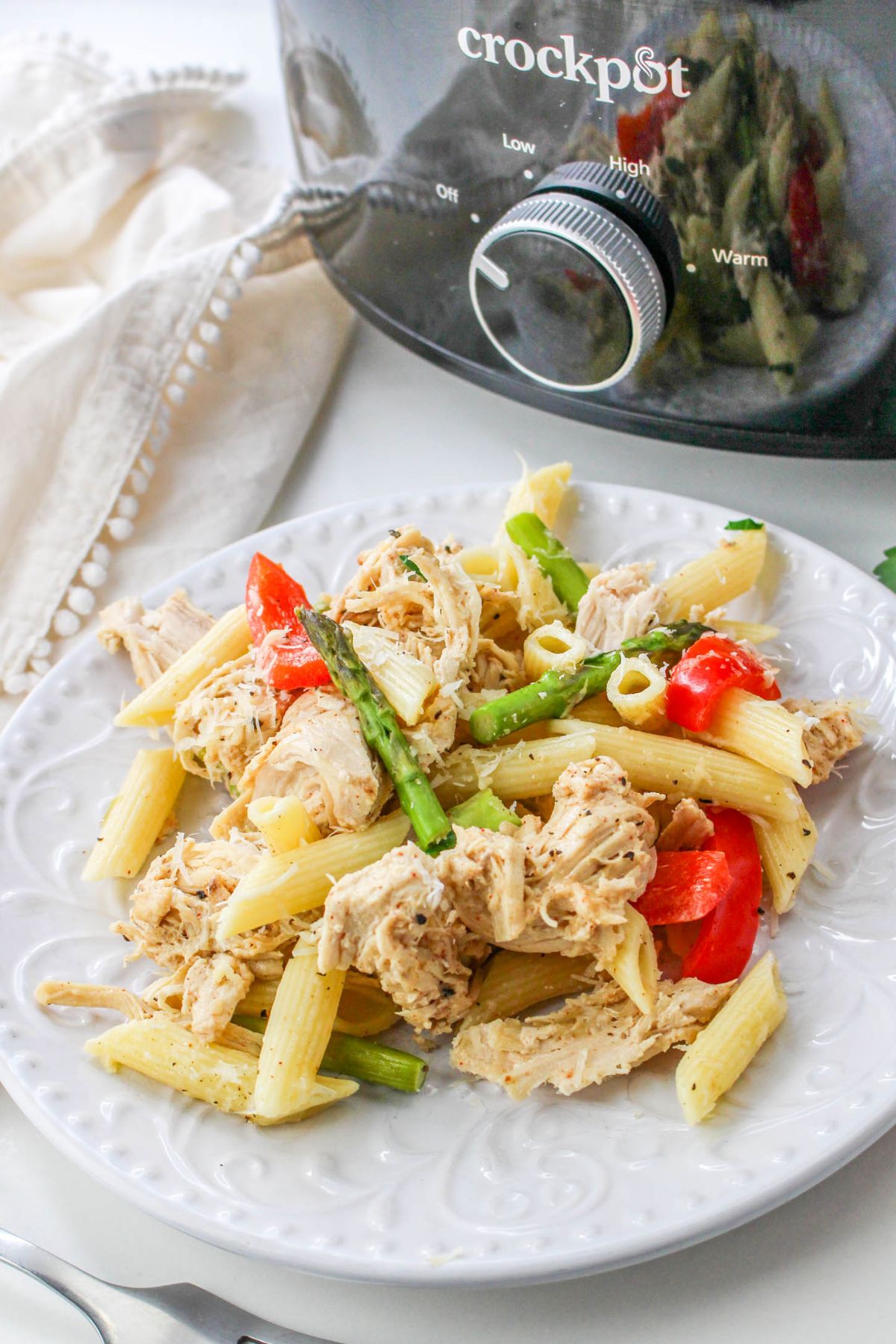 A plate of penne pasta with shredded chicken, asparagus, and red bell peppers sits in front of a Crockpot slow cooker.