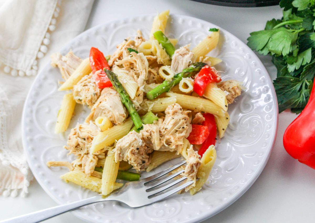 A white plate with penne pasta, shredded chicken, asparagus, and red bell peppers, with a fork resting on the plate.