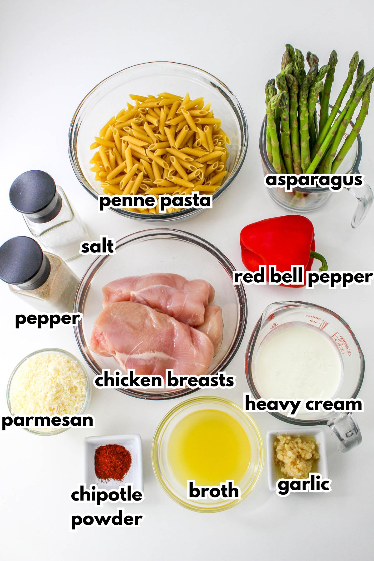 Top-down view of ingredients for a pasta dish: penne pasta, asparagus, red bell pepper, chicken breasts, heavy cream, broth, garlic, chipotle powder, parmesan cheese, salt, and pepper.