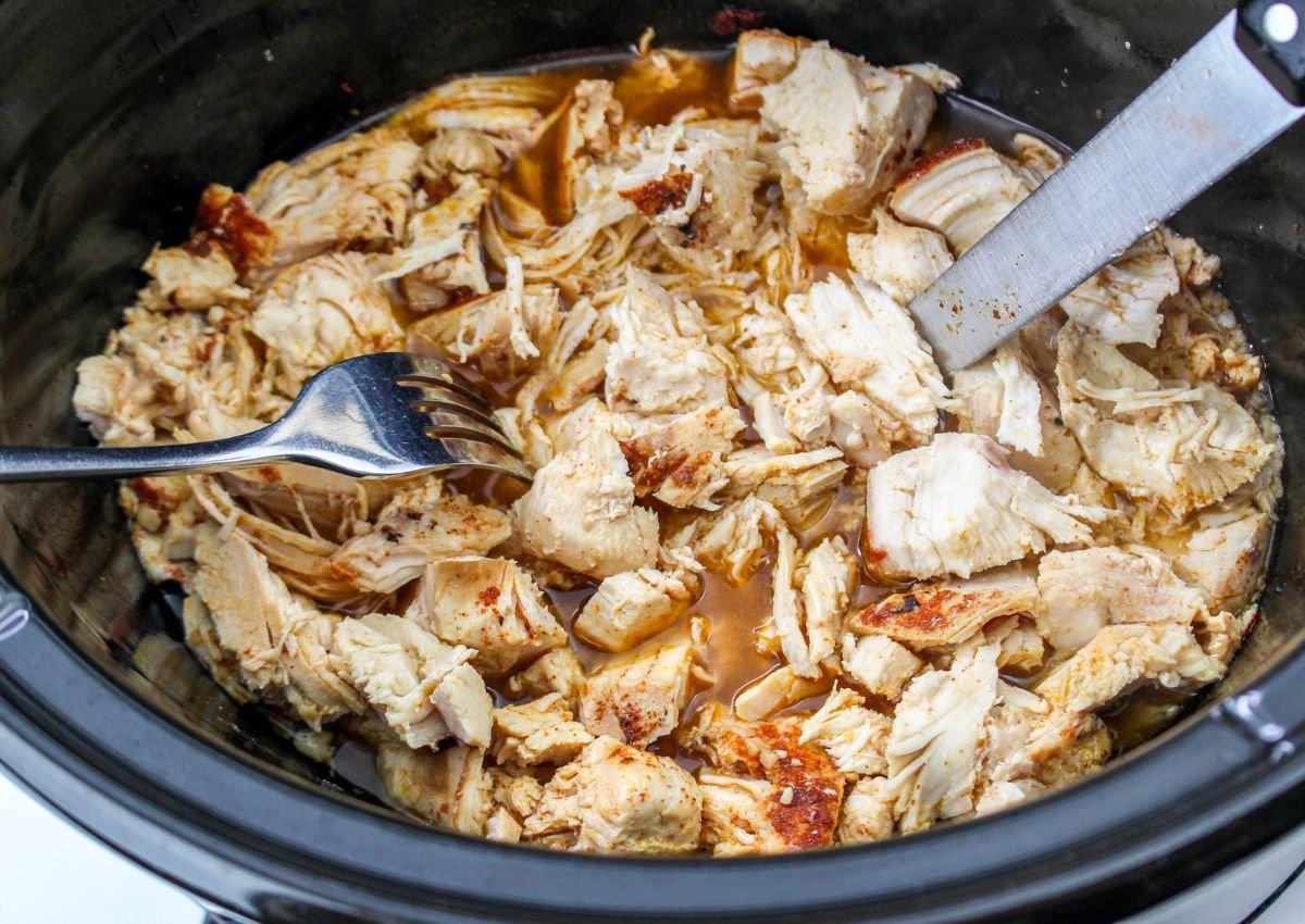 Cooked, shredded chicken breast in a slow cooker with a fork and knife resting on top of the meat.