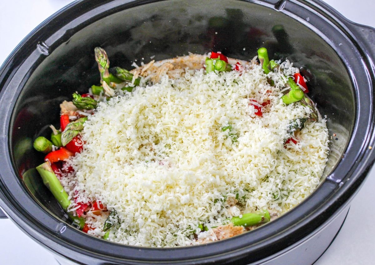 A slow cooker filled with shredded cheese, chopped asparagus, and red bell peppers on top of other ingredients.