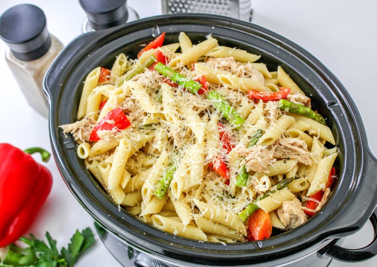 A slow cooker filled with penne pasta, shredded chicken, red bell peppers, green beans, and grated cheese, surrounded by salt and pepper shakers and a grater.