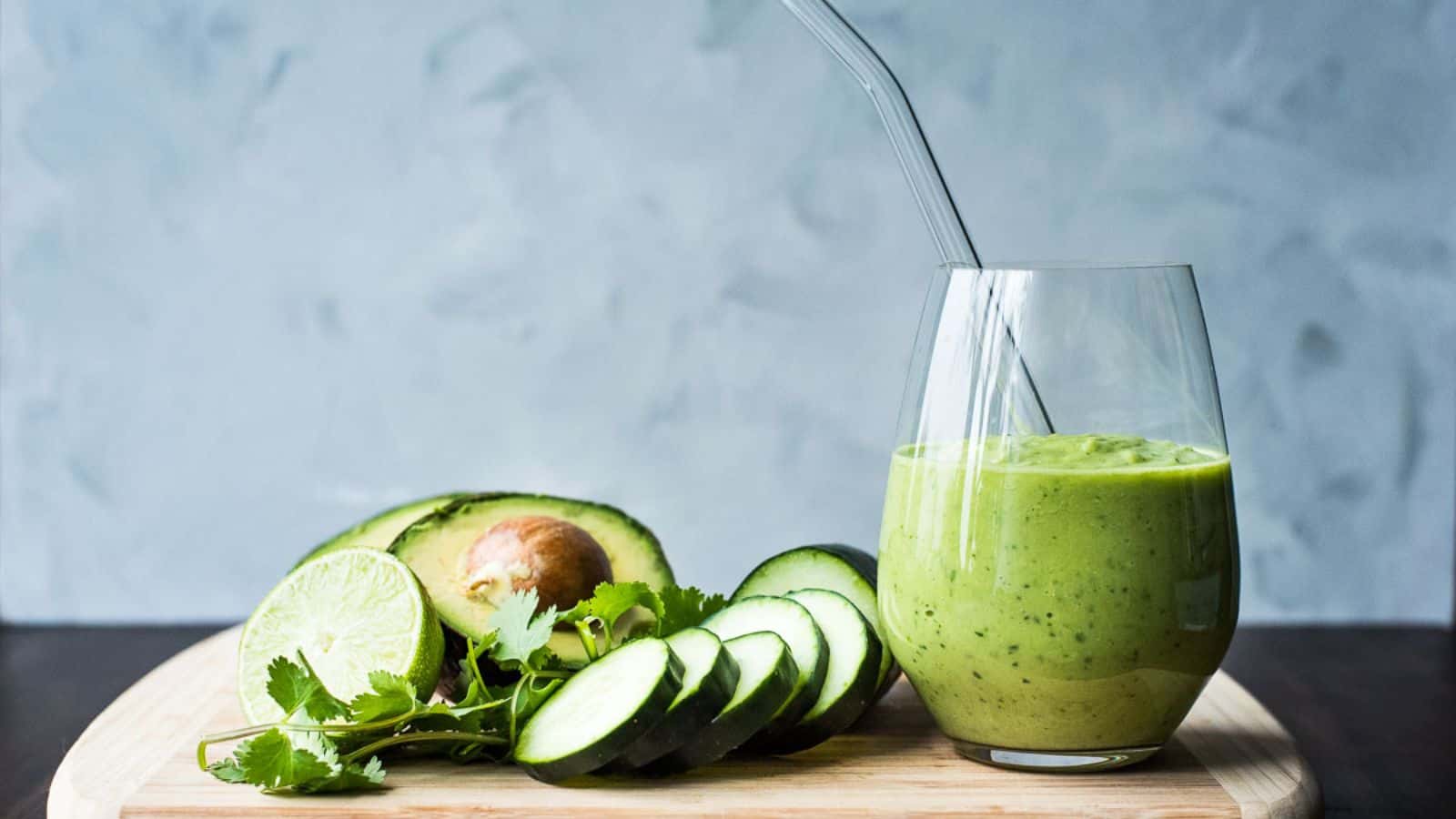 A glass of green smoothie with a straw sits on a wooden board beside sliced cucumber, avocado, lime, and cilantro.