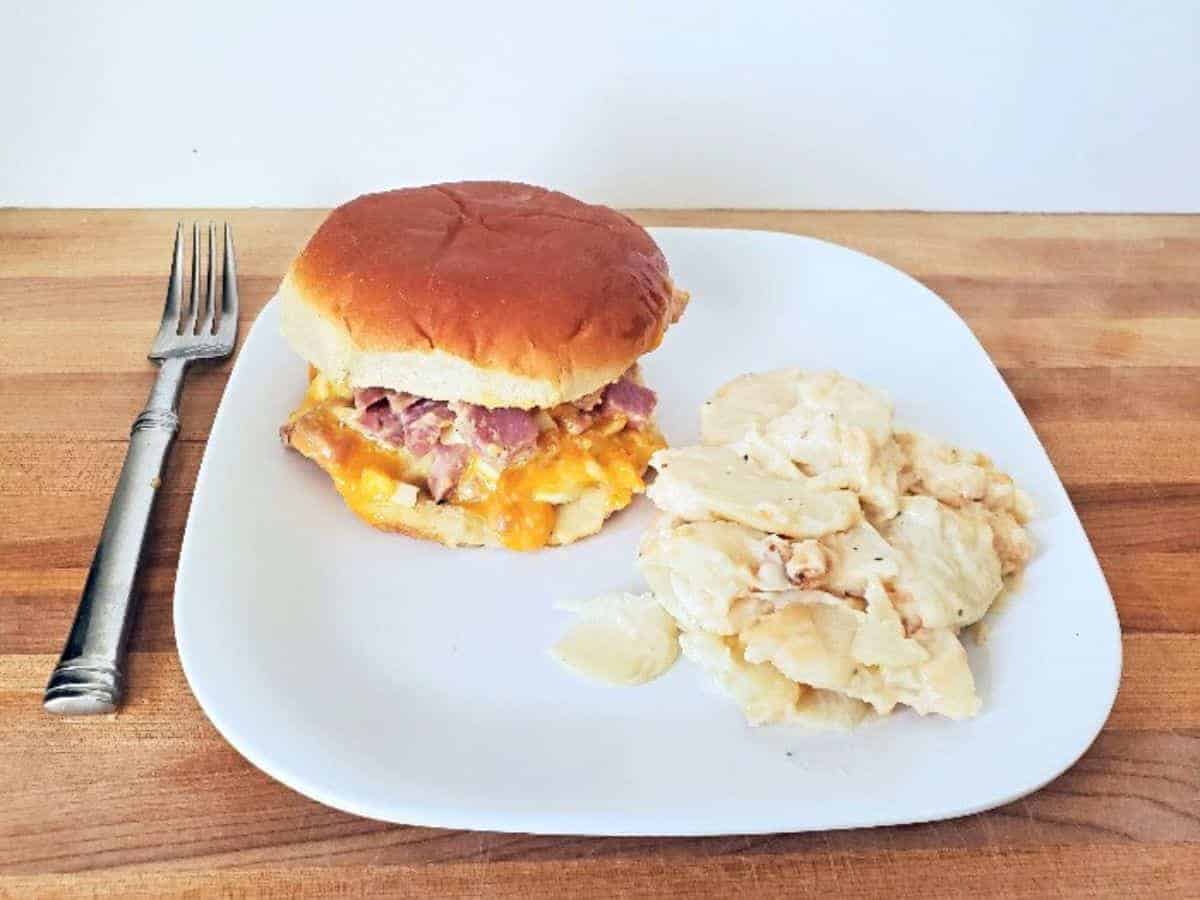 Image shows a white plate with a hot ham salad sandwich and scalloped potatoes.