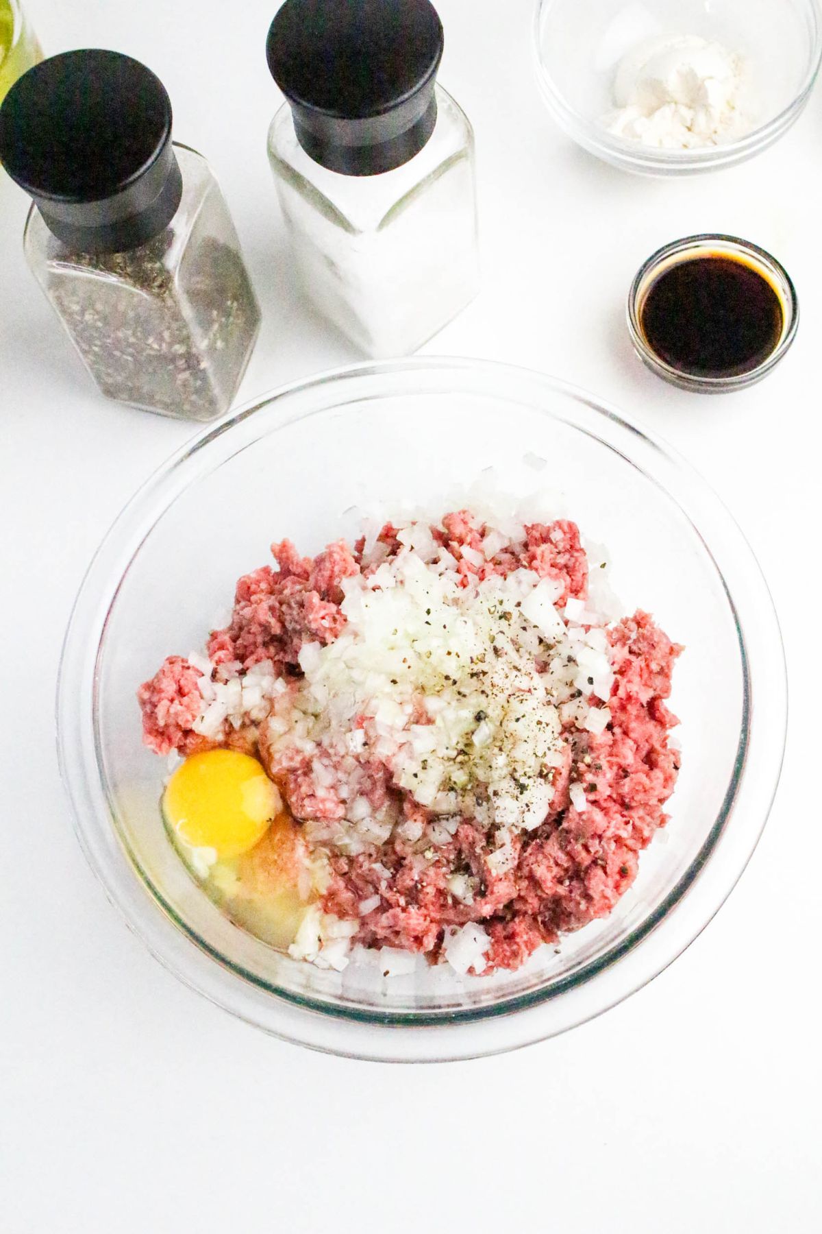 A glass bowl with ground beef, chopped onions, an egg, salt, and pepper. Nearby are salt and pepper shakers, a small bowl of sauce, and a dish with a white ingredient.