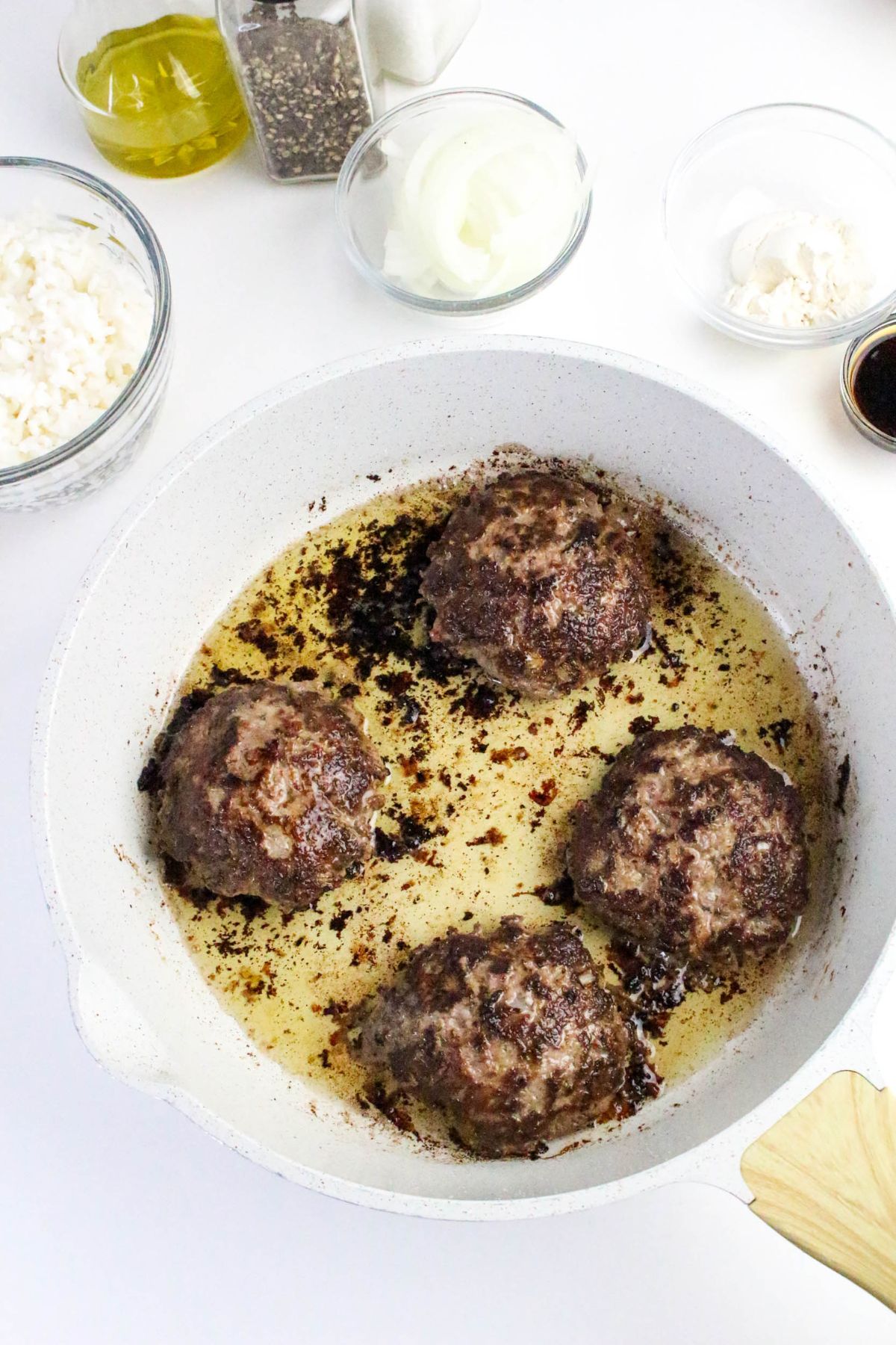 Four browned hamburger patties cooking in a white skillet, surrounded by bowls of ingredients including cottage cheese, onions, and seasonings.