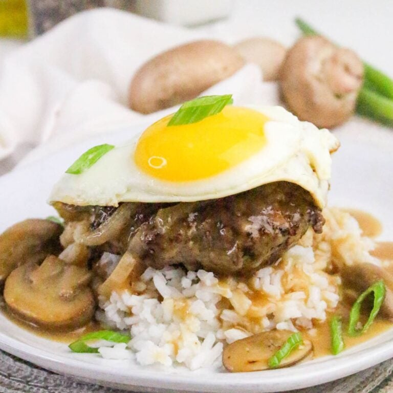 A hamburger patty topped with a sunny-side-up egg sits on white rice with brown gravy, sliced mushrooms, and green onions on a white plate.