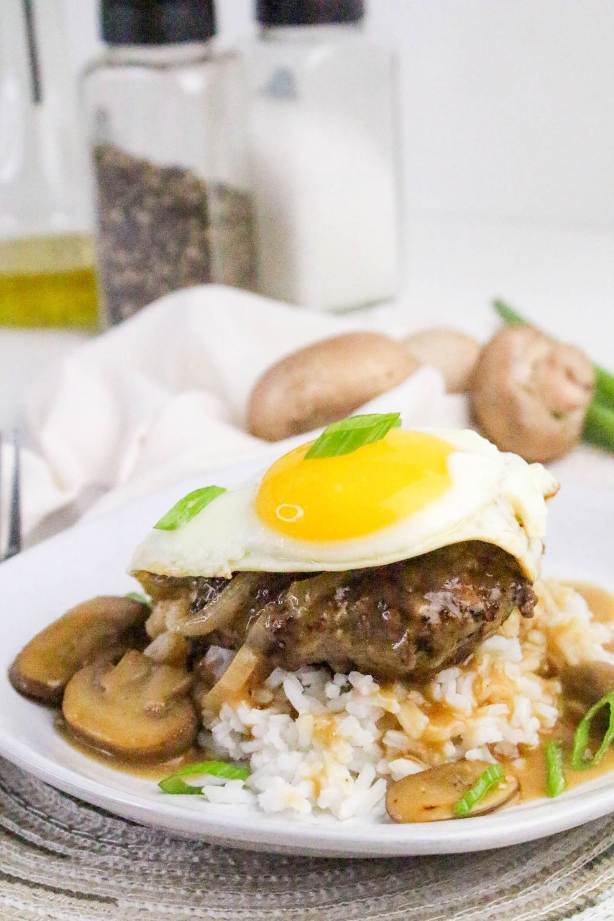 A plate of rice topped with a hamburger patty, sautéed onions, mushrooms, brown gravy, and a sunny-side-up egg, garnished with chopped green onions.