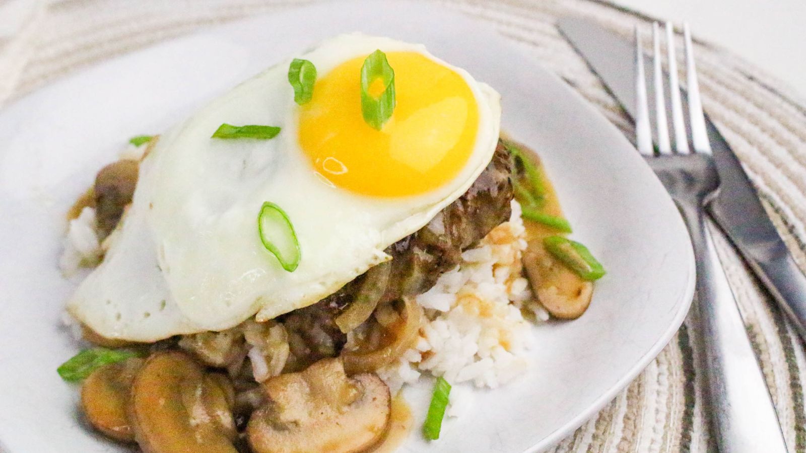 A plate of rice topped with sautéed mushrooms, a hamburger patty, brown gravy, and a sunny side up egg, garnished with sliced green onions. A fork and knife are beside the plate.