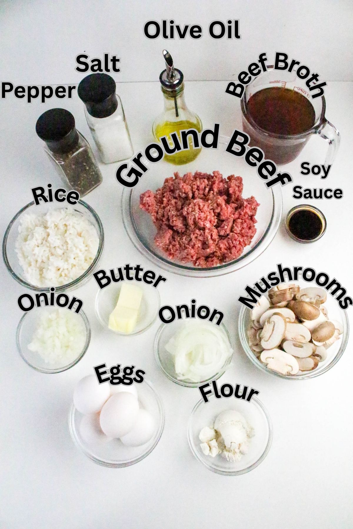 Ingredients for a ground beef dish arranged on a white surface, including ground beef, rice, onions, eggs, mushrooms, butter, flour, olive oil, beef broth, soy sauce, salt, and pepper.