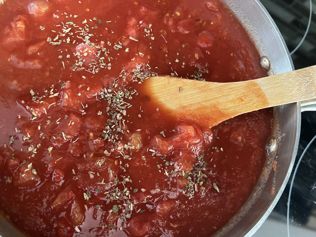 A pot of tomato sauce with visible tomato chunks and herbs, being stirred with a wooden spoon on a stovetop.