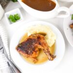 A plate with a roasted chicken leg quarter, mashed potatoes with gravy, and a gravy boat on the side.