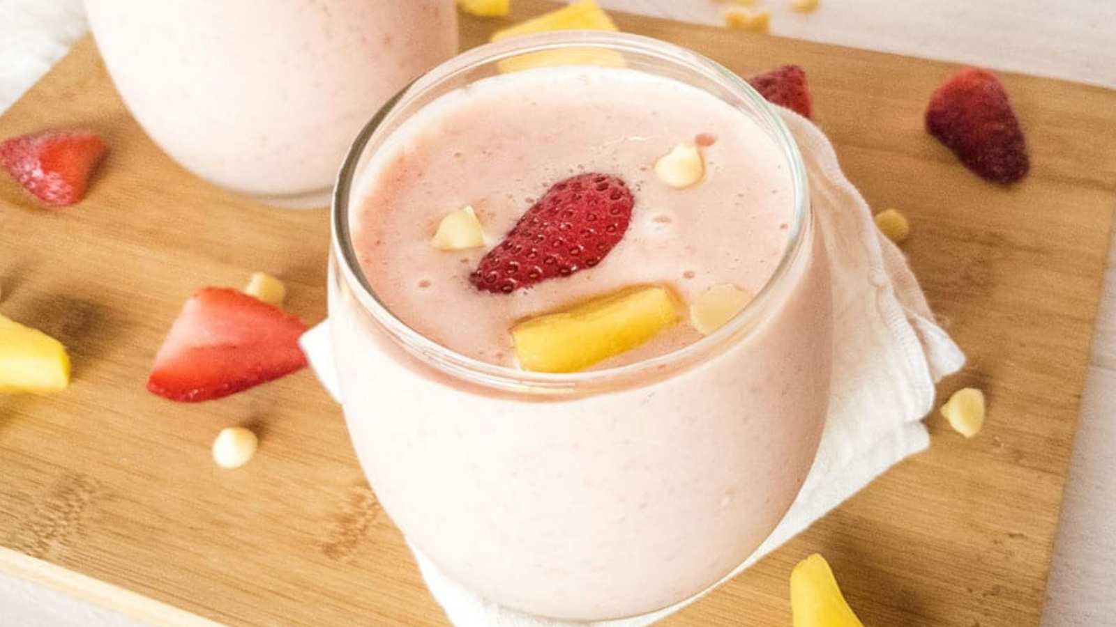 A glass of pink smoothie topped with strawberry slices, mango pieces, and white chocolate chips sits on a wooden board with more fruit scattered around.