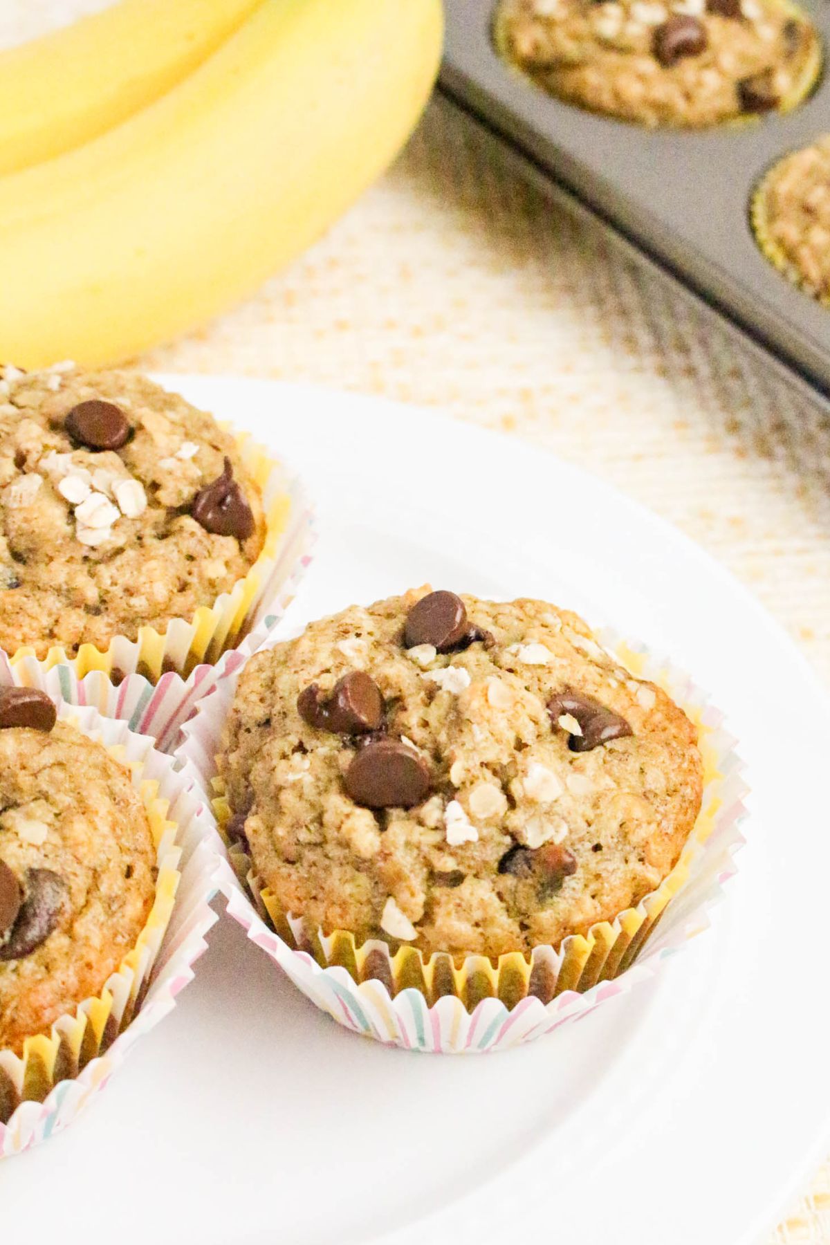 Three banana oat muffins with chocolate chips on a white plate, with a banana and muffin tray in the background.