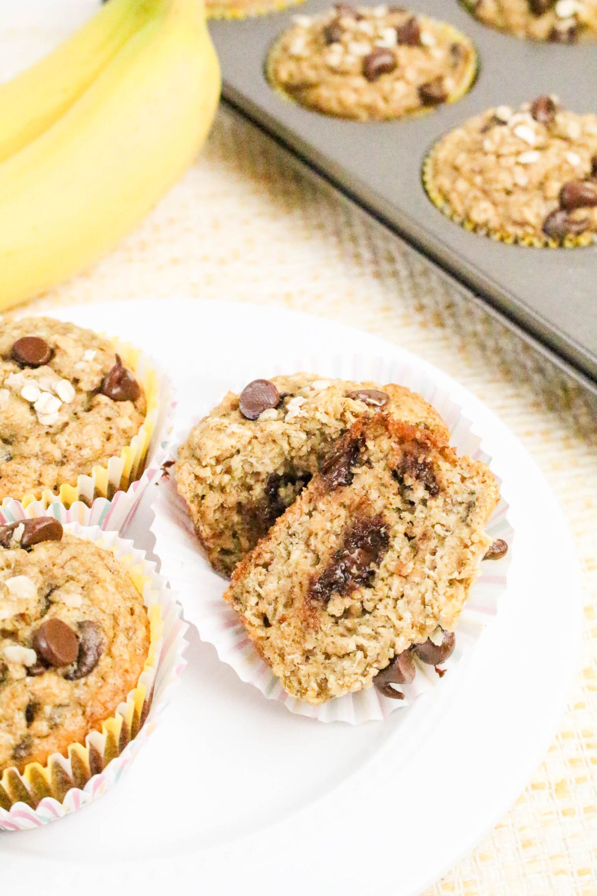 Banana chocolate chip muffins on a white plate, one muffin broken in half to show the inside; muffin tin and a banana in the background.