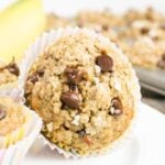 A close-up of a banana chocolate chip oat muffin in a paper liner, with more muffins in the background.
