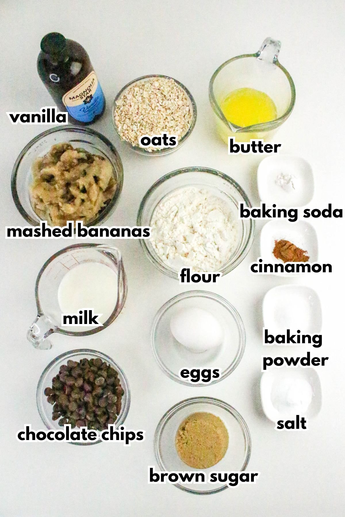 Top-down view of labeled baking ingredients in bowls and measuring cups, including mashed bananas, oats, flour, chocolate chips, eggs, milk, butter, vanilla, brown sugar, and spices.