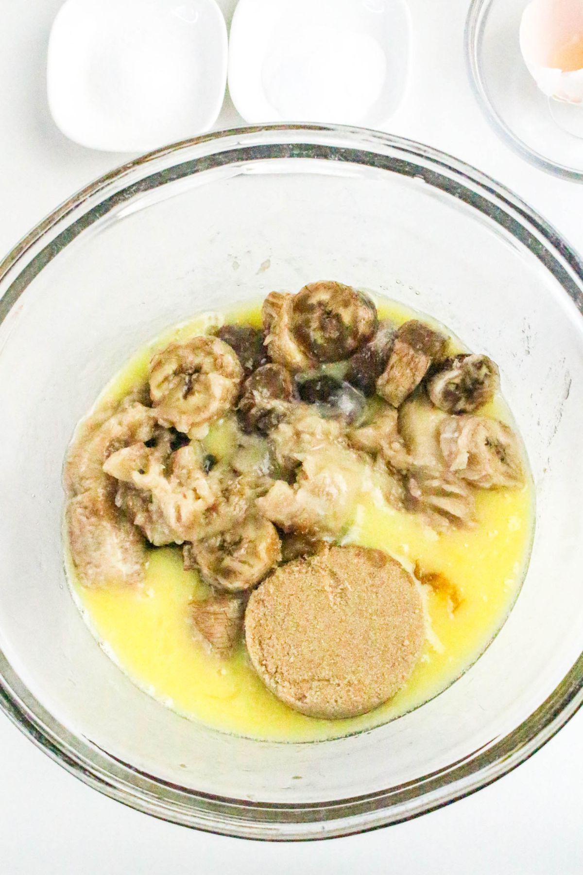 A glass bowl containing sliced bananas, melted butter, brown sugar, and other baking ingredients, with small bowls and an egg nearby.