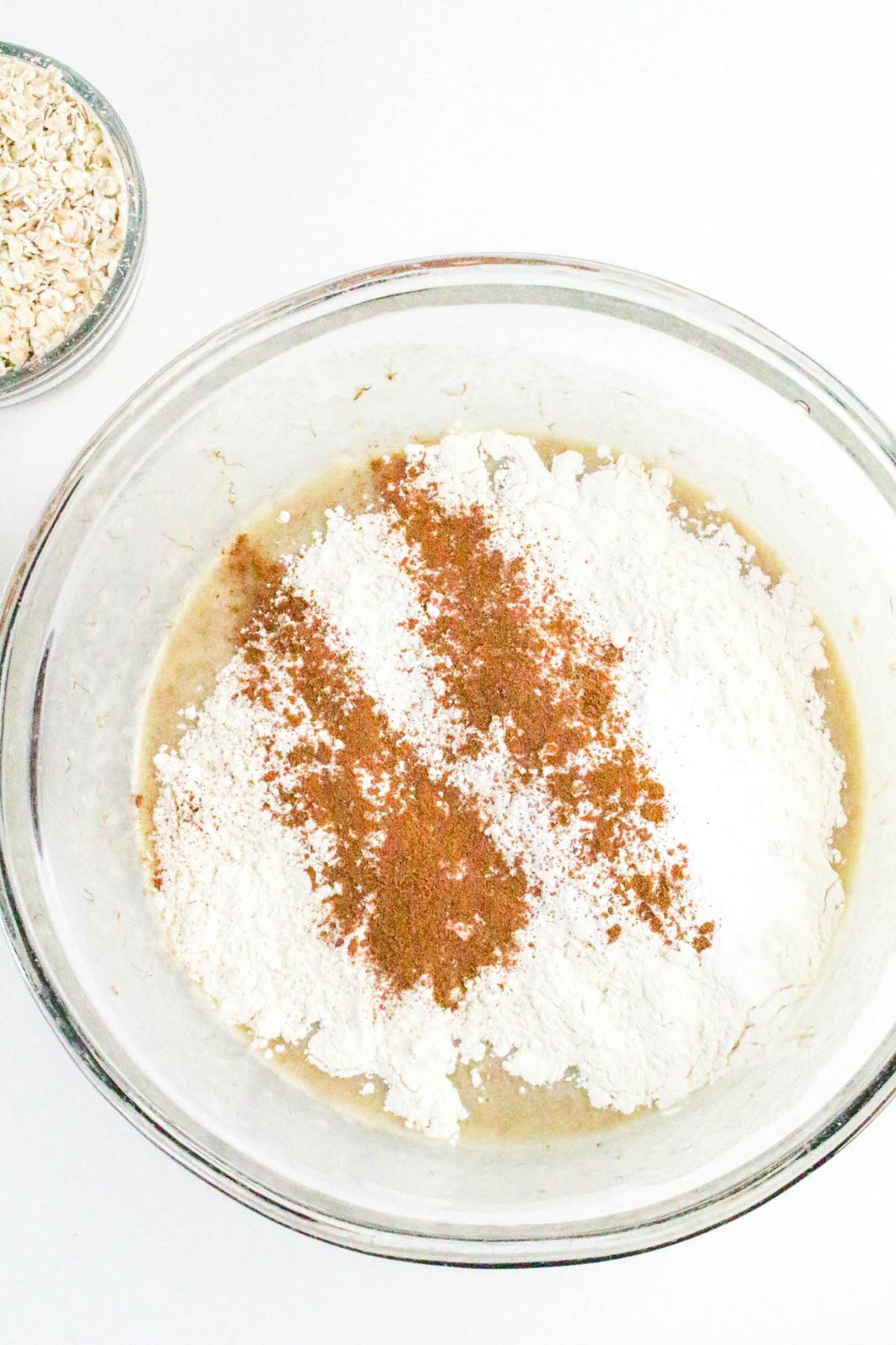 A glass mixing bowl with flour, baking powder, and spices on wet ingredients; a small bowl of oats is next to it on a white surface.