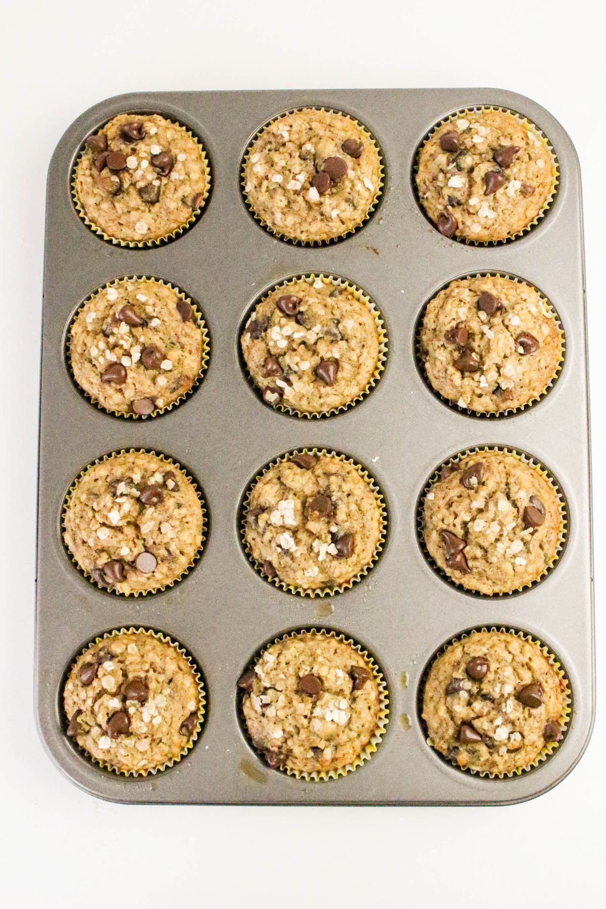 A muffin tin holds twelve baked oatmeal muffins topped with chocolate chips and oats, viewed from above on a white background.