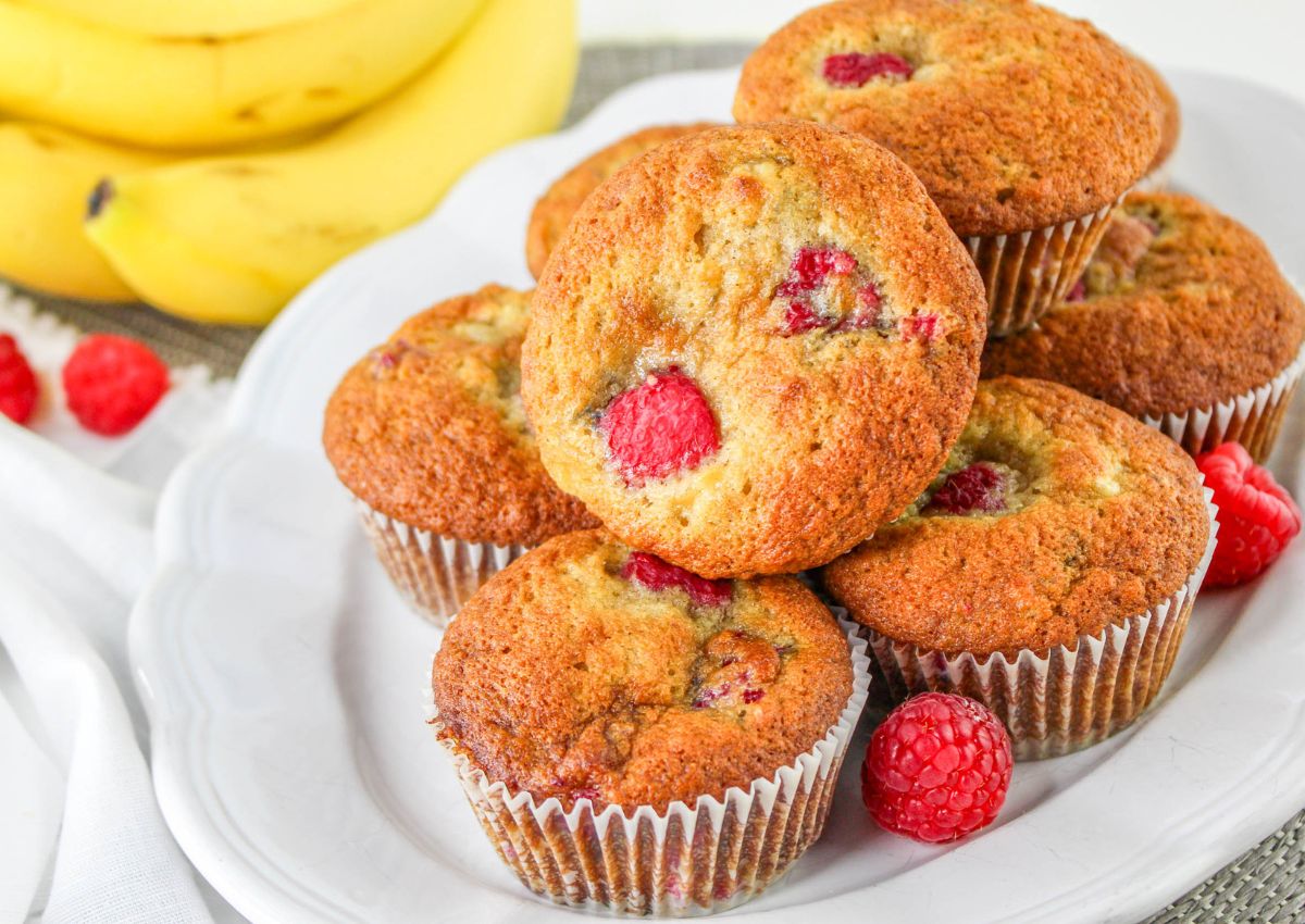 A white platter stacked with banana raspberry muffins with whole raspberries and bananas in the background.