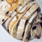 Sliced loaf of artisan bread with a golden crust and visible blueberries, arranged on a white plate.