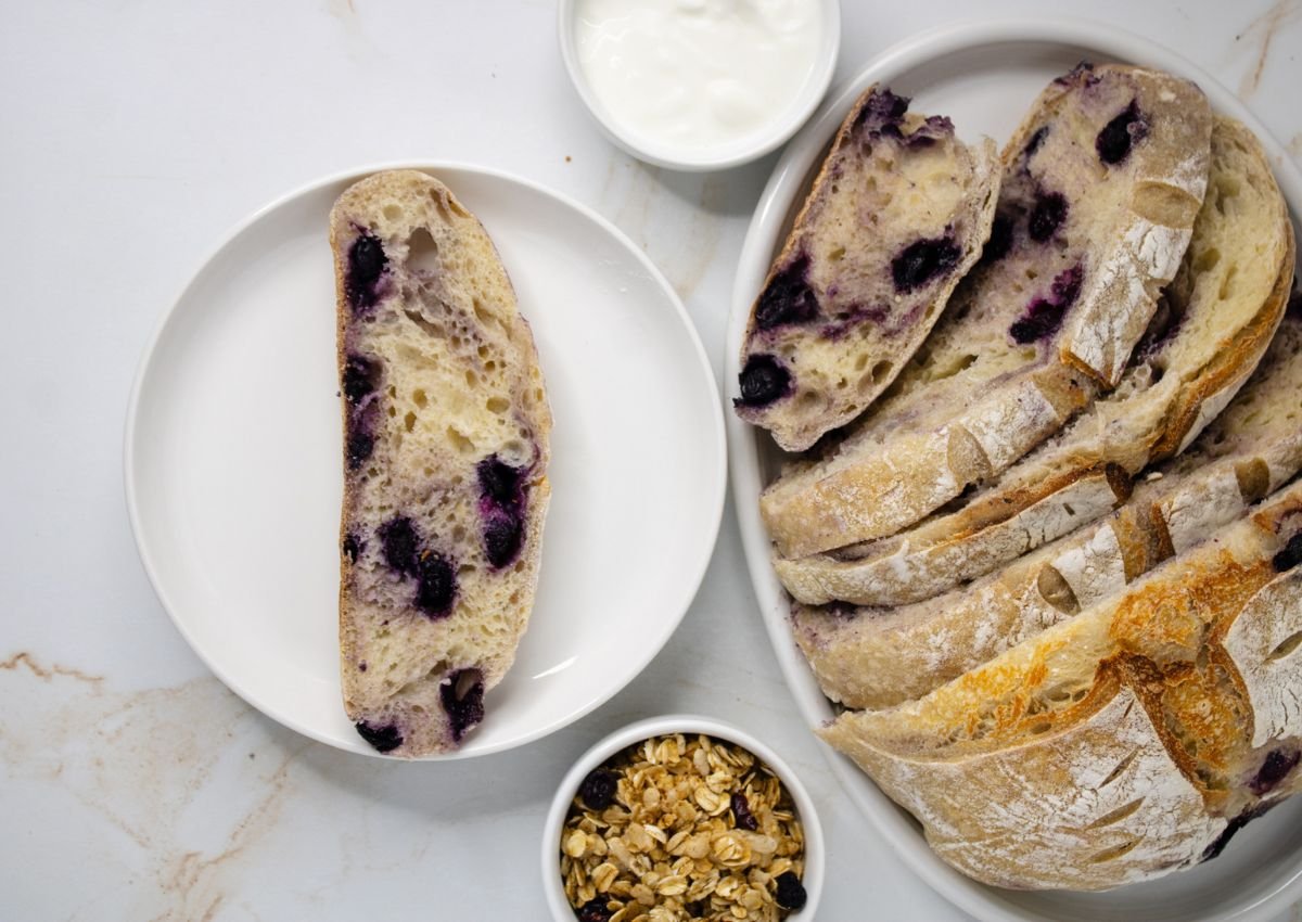 A sliced loaf of blueberry bread on a plate, with bowls of yogurt and granola on a white marble surface.
