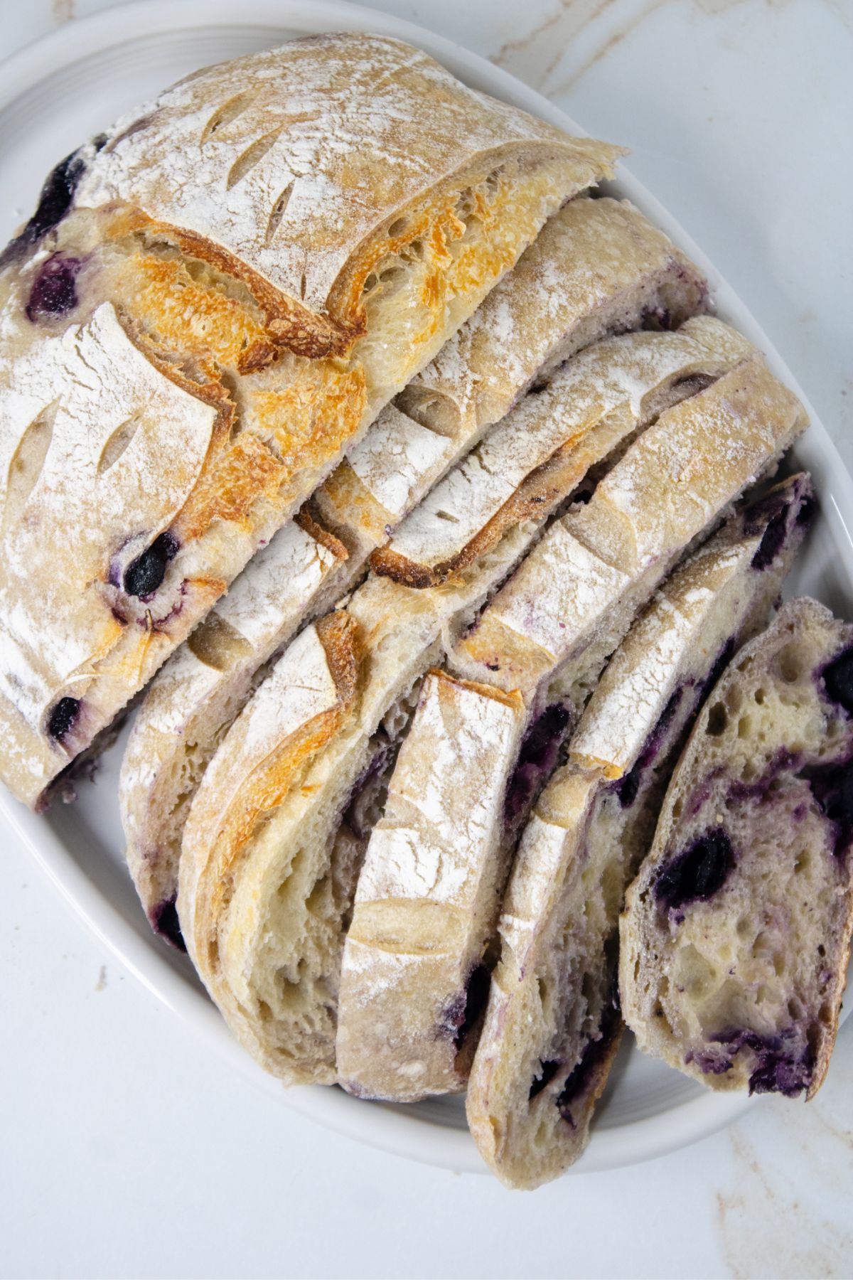 Sliced loaf of rustic bread with a dusting of flour and visible blueberries on a white oval plate.