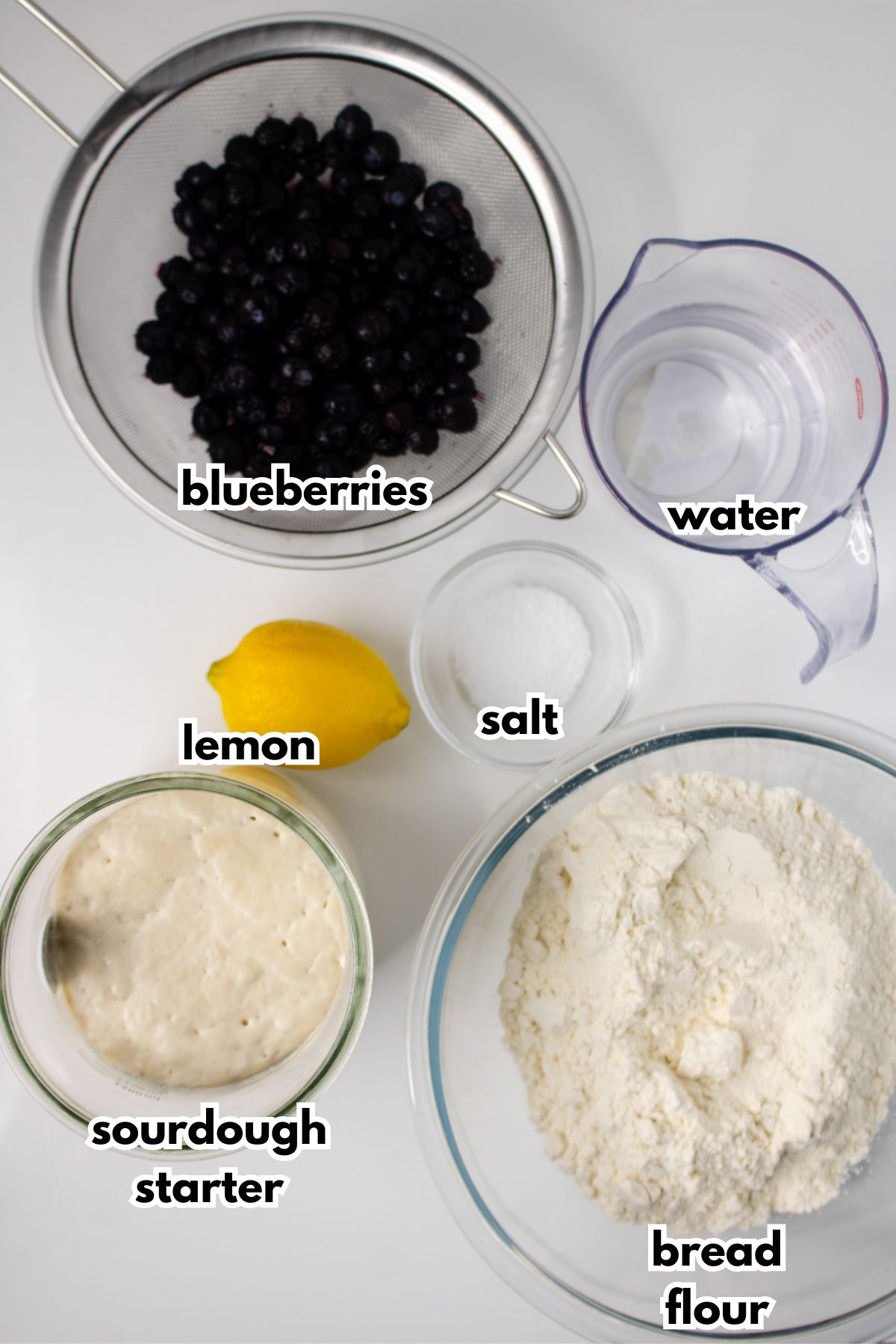 Top-down view of measured ingredients for baking: blueberries in a strainer, water in a jug, salt, a lemon, sourdough starter in a bowl, and bread flour in a mixing bowl.