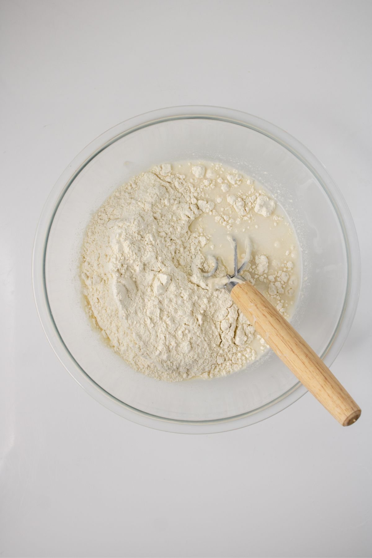 A clear glass bowl containing flour and liquid being mixed with a wooden-handled whisk, viewed from above on a white surface.